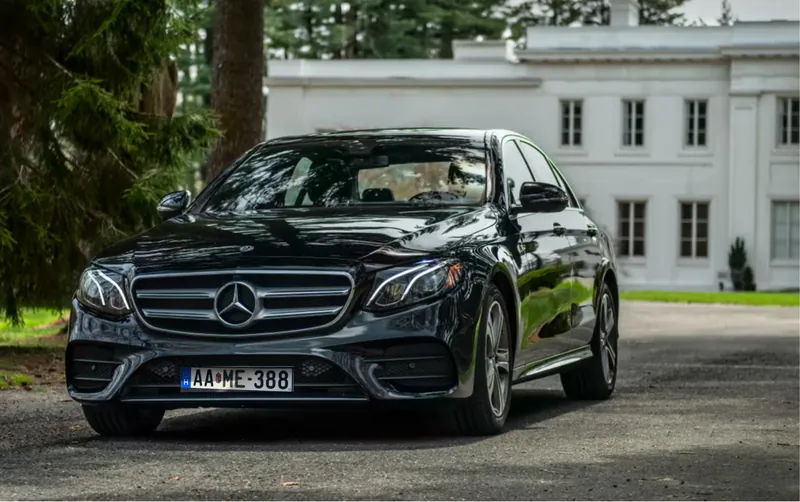 Fekete Mercedes-Benz E-osztály elölnézetből egy fehér kúria előtt, fák között